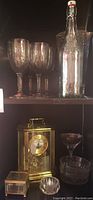Shelf display with four purple-tinted wine glasses, clear tall glass bottle with stopper, gold-tone mantel clock, and various glass items