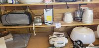Photo of wooden shelf with black and silver toaster, small metal lantern, white electric can opener, wrapped appliance, and multiple lamp shades.