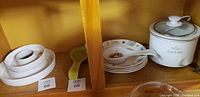 Shelf display of white crock pot, appetizer plates with matching spoon holders, two white ceramic ramekins with lids, and glass lid cover.