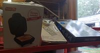 Photo showing boxed Dash pocket sandwich maker and two other boxed items including Pampered Chef mandolin and grill cover along with tissue holders on a shelf.