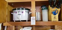 Shelf with large floral Crock-Pot, metal sifter, plastic pitcher with green lid, wooden Henckels cutlery holder with knives and scissors, white ceramic pitcher with colorful pattern below
