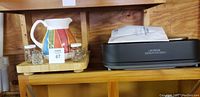 A wooden shelf holding the colorful Crate and Barrel pitcher and two small glass salt and pepper shakers on a wooden tray, next to the black Oster smokeless indoor grill with a manual on top.