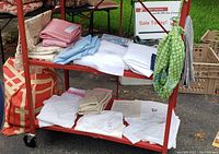 Two-level red metal cart holding various folded linens including pastel colored tablecloths, white linens, towels, and a hanging green floral shower curtain, with bags and crates nearby.