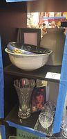 Wide shelf view of pottery bowls, jug, picture frame, and crystal vase with a boxed item and a decorative photo frame