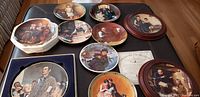 Over 10 Norman Rockwell collectible plates arranged on a table, featuring detailed artwork and various scenes, some plates stacked