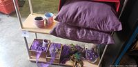Photo of two purple pillows, two mugs, purple basket, masquerade mask and other assorted decorations on shelf.