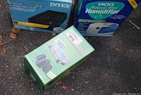 Photo showing boxed Intex air mattress, Vicks humidifier, and green box of bed risers on ground.