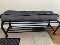 Metal shoe rack bench with cushioned seat in grey and black herringbone fabric. The seat is removable and the metal frame is black with horizontal bars for shoe storage.