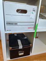 White storage box labeled as a storage box on a shelf with another black box beneath it, indicating box containers for items.