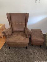 Brown faux suede wingback chair with matching ottoman shown on carpeted floor against a white wall, visible tufting on chair backrest, armrests and black legs.