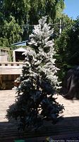 Full view of 90 inch artificial Christmas tree with white flocked branches, standing outdoors on wooden deck, showing overall tree shape and density.