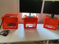 Three orange plastic crates on a table with computer monitors placed on top, visible window in background.