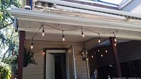 View of multiple clear glass bulbs attached to black wire string hanging from porch roof with some bulbs illuminated and one bulb appearing burnt out.