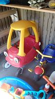 Side view of red and yellow Little Tikes toy car next to blue and yellow Kettler trike, stored in a shed with other toys.