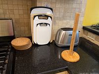 Four plastic cutting boards stacked upright with black handles, Stirling Oster toaster, wooden paper towel holder with round base, and six cork coasters stacked flat on kitchen counter.