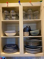 White cabinet with shelves holding stacks of plates, bowls, and glass jars with lids.