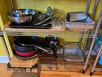 Photo showing frying pans and saucepan on lower shelf along with wooden cutting boards, metal wire mesh strainer, metal shredder, and tin foil containers.