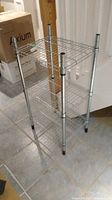 Three-tier chrome wire shelving unit shown angled, standing on tiled floor, next to cardboard boxes.