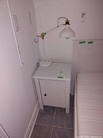 White wooden side table with a door and storage space inside, 17x14x25 inches, against a wall next to a white cushioned seat and accompanied by a white metal lamp with frosted glass shade.