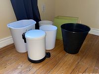 Set of six trash bins arranged on a wooden floor near a corner with a curtain, showing size, style, and color variety.