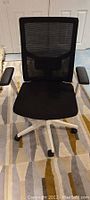 Front view of the black office chair showing mesh back with lumbar support and padded seat, white base, and armrests.