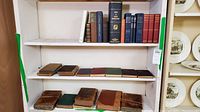 Books on two white shelves showing multiple hardcover books in leather and cloth binding with notable age wear.