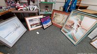 Photo showing five framed art pieces arranged on carpeted floor against wall