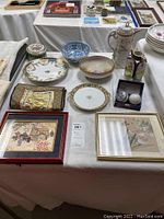Lot overview showcasing silk wall hanging, Noritake plate, teapot, decorative bowls, small vase, Chinese marble health balls, and framed art prints on display table.