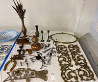 Wide view of all brass and mixed metal decorative items displayed on table, showing variety of metal art including mirror, feather holder, fish sculpture, horseshoe, candle holders, small vases, and wire knight silhouette.