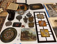 Overview of multi-cultural metal crafts including cast iron trivets and bookends, enamelware trays, enameled brass wall decor, and decorative metal items arranged on floor and table.