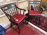 Pair of collegiate wooden armchairs with curved armrests, black and natural wood finishes, red seat cushions, and collegiate emblems on the backrests, sitting on a red patterned rug.