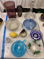 Overall shot of all items in the lot on a white tablecloth, including various glass vases, decanter, bowls, and decorative pieces.