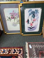 Two framed hummingbird illustrations angled upright on a carpeted floor with detailed rugs nearby, showing gold decorative frames and colorful matting.