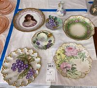 Top-down view of 7 porcelain china items including decorative plates and a small vase with floral and grapevine hand-painted designs and gold trim.