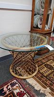 Full view of the round table showing glass top and woven rattan base on carpeted floor.