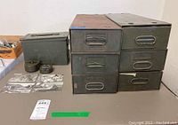 Photo shows six green metal military Safe-T-Stat storage boxes stacked in two columns, one military compass case, one olive drab ammunition box, and four black and white military postcards spread out on a table.