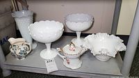 Overview of hobnail milk glass items including two footed compotes, ruffled bowl, Royal Tudor cream pitcher, and porcelain teapot placed on a shelf.