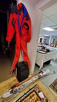Photo showing full view of red and blue adult ski suit hanging on a wall, white skis with bindings placed on the floor wrapped in plastic, and black wheeled suitcase underneath.