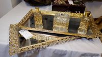 Two rectangular mirrored vanity trays with ornate golden filigree frames stacked, featuring three glass items with matching golden filigree detail arranged on the top tray.