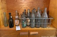 Front view of 16 glass vintage beverage bottles, mostly dusty, in a metal wire rack, along with some loose bottles on the side.