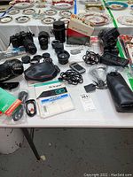 Vintage cameras, lenses, accessories, film boxes, and manuals spread on table with decorative plates in background.