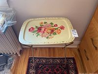 Single vintage metal TV tray with cream background and colorful floral artwork, supported on metal foldable legs, shown standing on a patterned carpet.