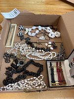 Top-down view of box showing assorted costume jewellery including white and black necklaces, earrings, and watch bands in separate compartment.