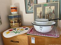 Overview of the enamel bowls, trays, placemats, and tins arranged on a wooden cabinet with paintings behind