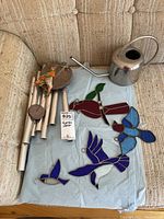 All items arranged on a light blue cloth on a beige couch showing the wind chime with metal tubes, three stained glass bird wall hangings in red, blue, and purple, and a small silver watering can.