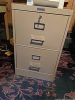 Front view of the beige metal file cabinet showing two drawers each with label holder and central metal handle; lock key inserted in top drawer.