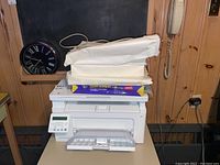 White HP LaserJet Pro printer with digital control panel, trays open, power cord, and multiple paper reams stacked on top.