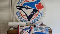 Large round Toronto Blue Jays cardboard poster on top of a dresser along with other rolled Toronto sports posters and pennants.