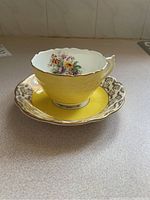 Photo showing yellow and white scalloped teacup with floral interior and gold trim, paired with matching saucer with gold patterned rim and yellow center.