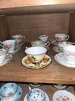 Shelf holding seven assorted teacups and matching saucers with various floral and gold designs from different brands.